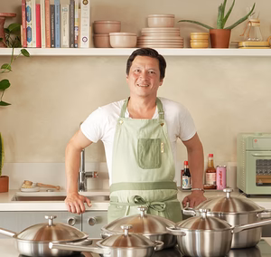 Man in a kitchen wearing a green apron with pots and pans in front of him.