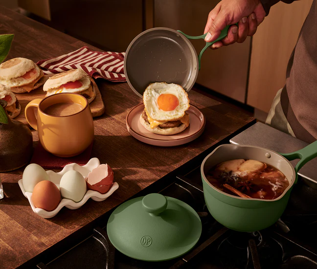 A person places a fried egg onto an English muffin sandwich, with mugs, eggs, and a saucepan of tea or broth on a wooden kitchen counter.